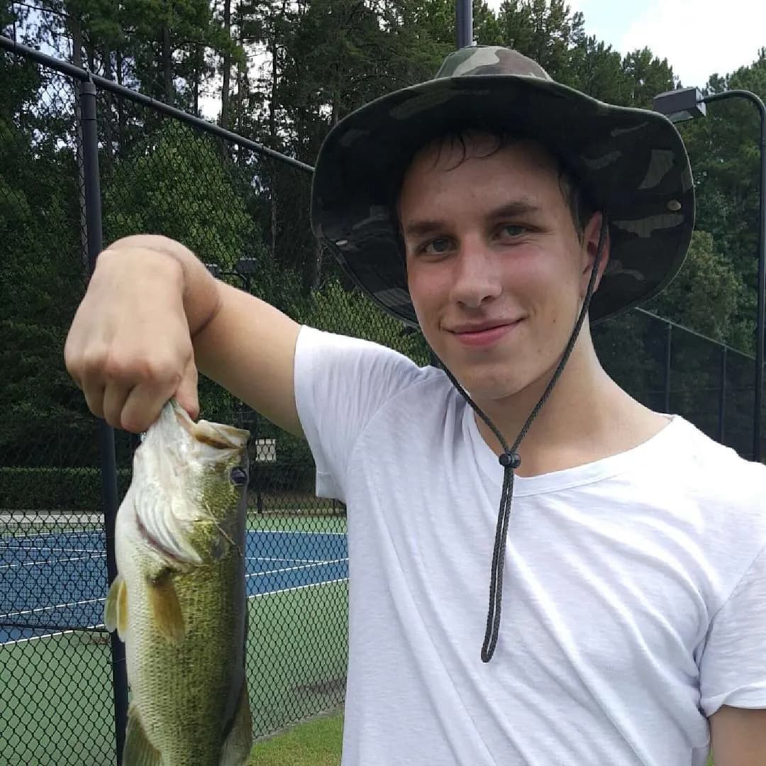 Tyler Henn holding a largemouth bass beside an outdoor tennis court